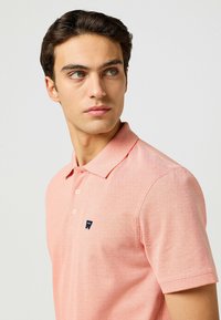 Polo shirt rosa chiaro con maniche corte, tessuto strutturato, patta con tre bottoni e un piccolo logo del marchio blu navy sulla parte sinistra del petto.