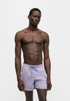 Muž tmavej pokožky bez košele, ktorý má na sebe svetlofialové plavecké šortky s modrým šnúrkom, stojí na jednoliatom bielom pozadí.