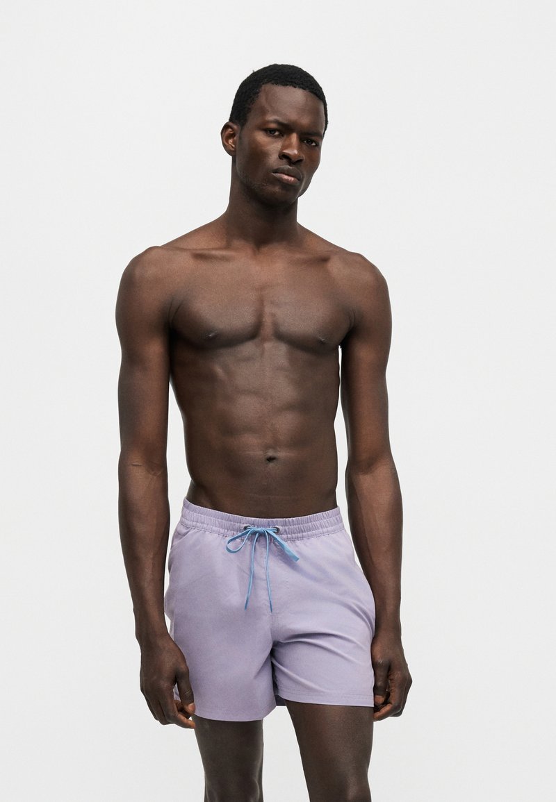 Mężczyzna o ciemnej karnacji bez koszulki, ubrany w jasnofioletowe kąpielówki z niebieskim sznurkiem, stojący na jednolitym białym tle.