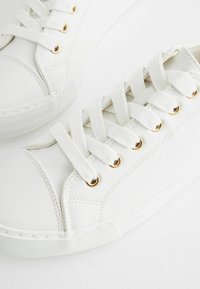 Vita sneakers med en slät överdel i syntetläder, gyllene ögla och platta bomullssnören. Strukturerad vit gummisula.