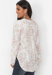 Blusa floreale in tessuto leggero, con uno sfondo bianco decorato da fiori rosa e gialli, maniche lunghe e un orlo leggermente arrotondato.