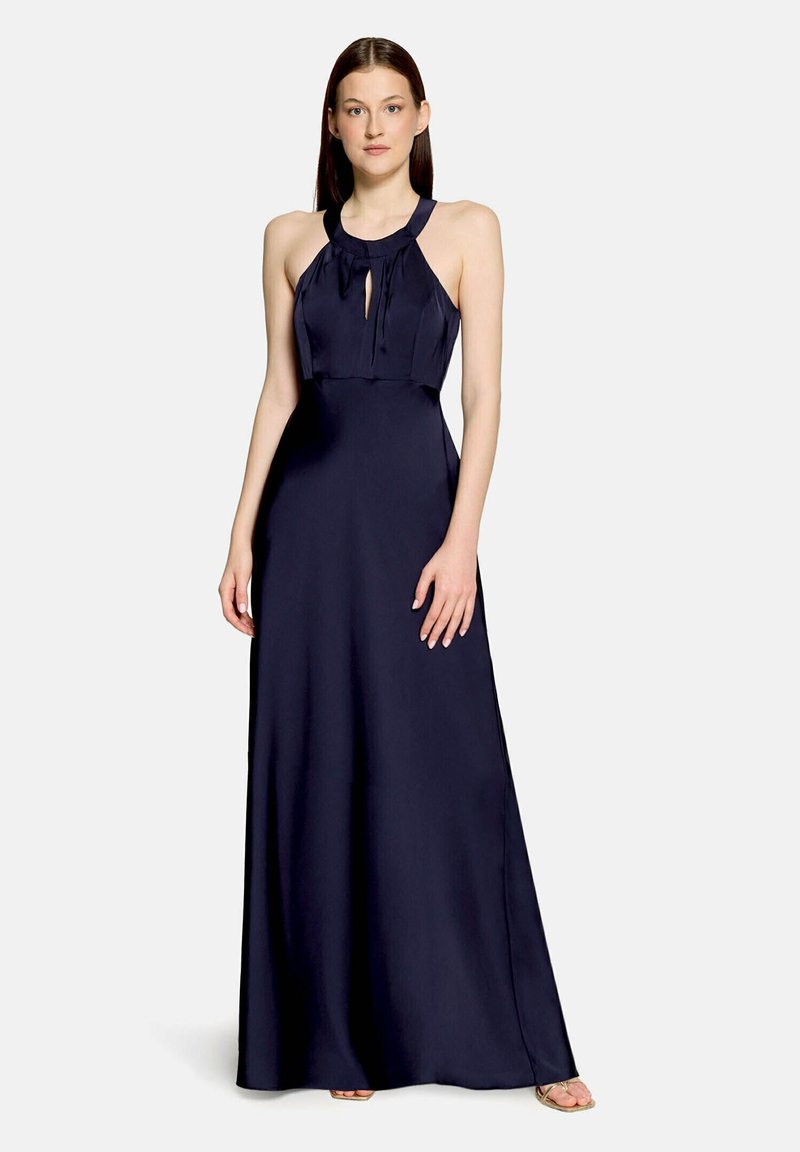 Femme portant une longue robe dos nu bleu marine avec un détail en forme de trou sur le devant, debout les mains détendues le long du corps.