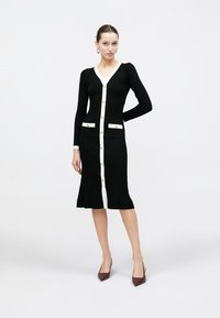 Μαύρο ριμπ φόρεμα με μακριά μανίκια, λευκό εμπρόσθιο μέρος με κουμπιά και δύο εμπρόσθιες τσέπες. Ολοκληρωμένο με χρυσούς κουμπιά και μήκος μέχρι το γόνατο.