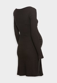 Přiléhavé černé těhotenské šaty s dlouhým rukávem a ribbed vzorem, z profilu viditelný těhotenský pupík, na bílém pozadí.