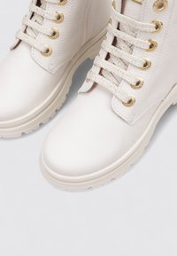Bottes en cuir couleur crème avec des semelles épaisses, œillets dorés et lacets assortis bien serrés, sur un fond clair.
