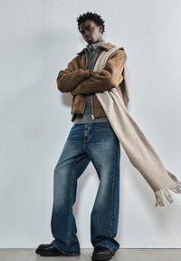 Mladenič oblečen v rjav usnjeni jakni, sivo pulover, široke modre hlače, črne čevlje in dolgo bež šal, ki mu pada čez eno ramo.