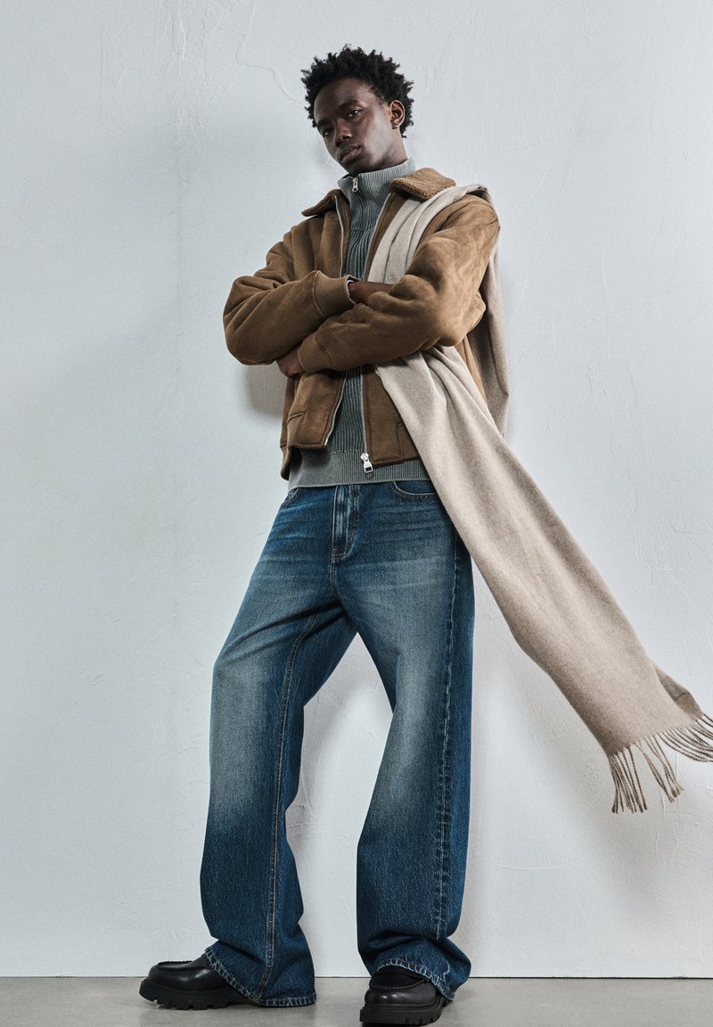 Mladenič oblečen v rjav usnjeni jakni, sivo pulover, široke modre hlače, črne čevlje in dolgo bež šal, ki mu pada čez eno ramo.