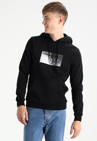 Zwarte hoodie van katoenmix met een voorkant kangaroo pocket, voorzien van een monochroom grafisch ontwerp op de borst. Blauwe spijkerbroek completeert de outfit.