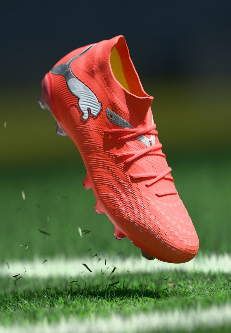 Botín de fútbol naranja brillante con superficie texturizada, detalles en plateado y tachones negros; suspendido sobre césped con pequeños restos.