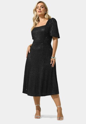 Femme portant une robe midi noire scintillante avec des manches ballon, des sandales argentées, un bracelet argenté et des boucles d'oreilles, debout devant un fond uni.