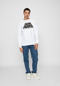 Hvid langærmet t-shirt med sort "STAR WARS" grafikprint, parret med blå denim cargo-bukser og hvide sneakers.