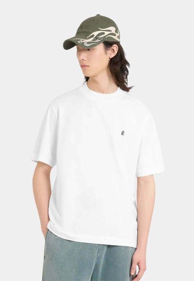 Jeune personne aux cheveux longs et foncés portant un t-shirt blanc surdimensionné et une casquette vert olive avec un motif de flammes blanches, regardant sur le côté.
