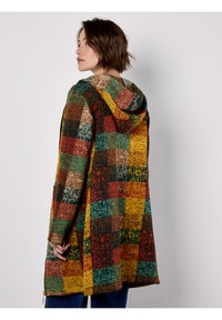 Długi kardigan z kapturem w kratę, z wielokolorowymi kwadratami w kolorach czerwonym, zielonym, żółtym i brązowym, z bocznymi kieszeniami i luźnym krojem.