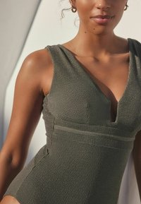 Olive green one-piece swimsuit with a textured pattern, deep V-neck, thick straps, and a fitted silhouette. Accented with subtle detailing.