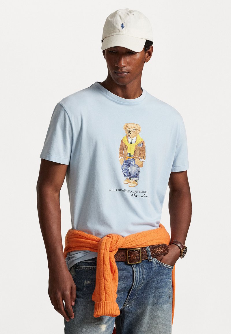 Camiseta azul-clara com um gráfico de um urso vestido com um casaco castanho, um lenço amarelo e um fato de ganga, combinado com um suéter laranja amarrado à cintura.