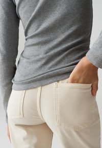 Persona che indossa pantaloni beige e una maglia grigia a maniche lunghe con la mano posata nella tasca posteriore dei pantaloni.