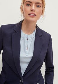 Blazer navy sartoriale con rever a lancia, due bottoni e tasche frontali, abbinato a una blusa a righe blu chiaro con dettaglio a occhio di bue.