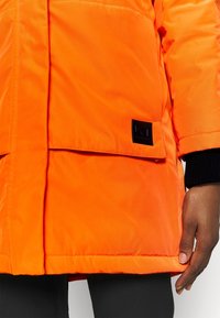 Gros plan sur la main d'une personne reposant près de la poche d'un manteau orange vif avec un écusson logo noir.