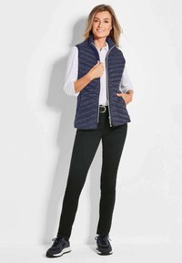 Marineblauer Steppweste mit Reißverschluss vorne, stehendem Kragen und horizontalen Steppdetails; kombiniert mit einem weißen Hemd und schwarzen Hosen, dazu dunkle Sneaker.