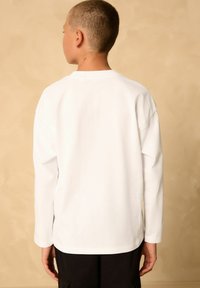 Valkoinen pitkähihainen t-paita, jossa on rento istuvuus ja pyöreä kaula-aukko. Ominaisuuksiin kuuluu suora helma ja hihansuut, valmistettu pehmeästä puuvillamateriaalista.