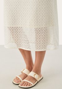 Robe en dentelle blanche avec des motifs floraux élaborés, associée à des sandales beige à large bride et détail de boucle circulaire.