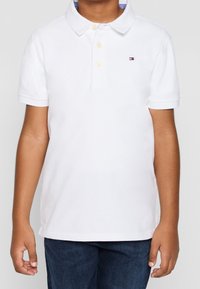 Junge trägt ein weißes Poloshirt mit drei Knöpfen und einem kleinen rot-weiß-blauen Logo auf der linken Brust, kombiniert mit dunkelblauen Jeans.