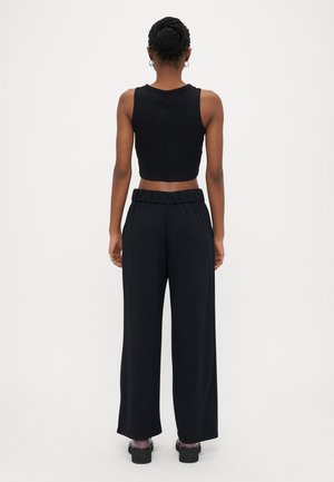 Kvinde med fletninger, iført sort ærmeløs crop top, sort vidde-højtaljede bukser og sorte sko, stående med ryggen mod en hvid baggrund.