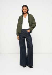 Casaco verde oliva com um corte descontraído e bolsos, combinado com jeans de perna larga azul escuro. O tecido parece liso com costura visível.