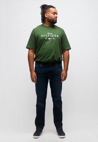 Άνδρας με ντραντρέξ φοράει πράσινο T-shirt Tommy Hilfiger, σκούρο τζιν και μαύρα αθλητικά παπούτσια, στέκεται μπροστά σε απλό λευκό φόντο.