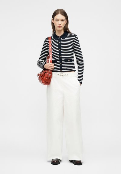 Femme portant un cardigan à boutons rayé noir et blanc, un pantalon large blanc, des chaussures noires, et portant un sac à bandoulière rouge.