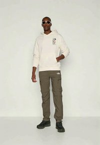 Férfi fehér kapucnis pulcsit, olíva színű cargo nadrágot, fekete túracipőt és fehér napszemüveget visel, egy sima világos háttér előtt áll.