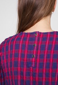 Baumwollbluse mit lila und rotem karierter Musterung, ausgestattet mit drei silbernen Knöpfen auf der Rückseite und einem runden Ausschnitt. Strukturierter Stoff.