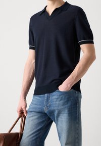 Polo à manches courtes bleu marine avec un col en V, agrémenté d'accents blancs aux poignets et d'un ourlet côtelé. Porté avec un jean bleu clair et un sac marron.