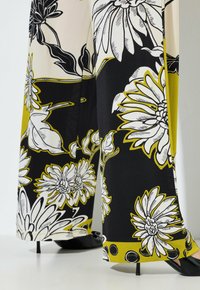 Spodnie o szerokich nogawkach z dużym czarnym, białym i żółtym kwiatowym nadrukiem, noszone z czarnymi butami na wysokim obcasie na białej podłodze.