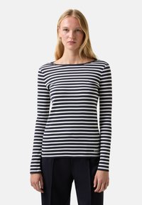 Young woman with long blonde hair wearing a black and white horizontal striped long-sleeve shirt and black pants, standing against a plain background.