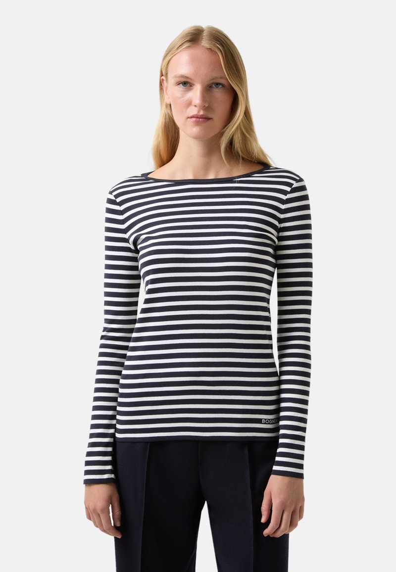 Young woman with long blonde hair wearing a black and white horizontal striped long-sleeve shirt and black pants, standing against a plain background.