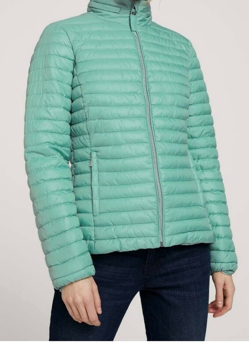 Femme portant une doudoune matelassée bleu sarcelle clair avec une fermeture éclair frontale et des poches latérales zippées, associée à un jean bleu foncé.