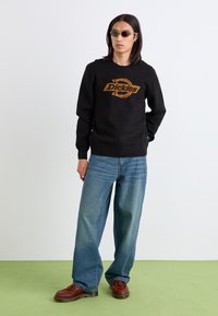 Schwarzer Strickpullover mit orangefarbenem "Dickies"-Logo, kombiniert mit hellblauen weitem Jeans und braunen Schuhen. Modell trägt Sonnenbrille.