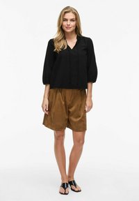 Blusa nera, in fit morbido, con scollatura a V e dettagli con cordino abbinata a pantaloni corti marroni con pieghe. Il modello indossa sandali neri.