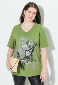 Camiseta verde de manga corta con escote en V, con un gráfico de un ciervo y acentos artísticos en negro y gris. Combinada con un clutch de tejido.