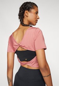 Rosa kortärmad topp med knutbakdesign som avslöjar ett svart texturerat plagg under. Modellen bär högmidjade svarta leggings.