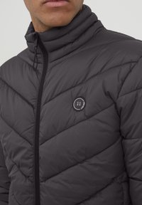 Jachetă neagră cu umplutură, cu guler înalt, model în zigzag, închidere cu fermoar și un logo rotund cu numărul 11 în partea stângă.