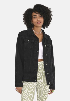 Jonge vrouw met krullend haar die een zwarte spijkerjas, een wit crop top en groene broek met patroon draagt, staand tegen een effen achtergrond.
