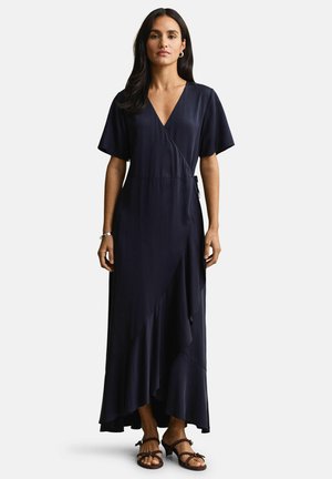 Vrouw draagt een donkerblauwe maxi-jurk in wikkelstijl met korte mouwen en een gerimpelde zoom, gecombineerd met bruine sandalen met hak en minimalistische sieraden.