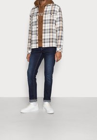 Checkered flannel jacket with front pockets in beige, brown, and white; layered over a brown hoodie. Dark denim jeans and white sneakers.