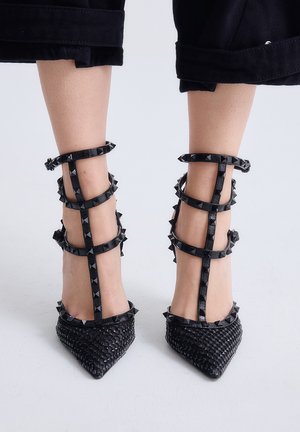 Tacones negros tejidos con punta estrecha y múltiples tiras con tachuelas en el tobillo, combinados con pantalones cortos negros sobre un fondo liso.