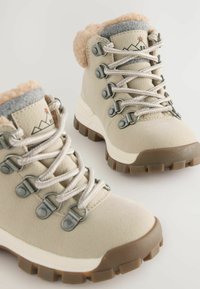 Beige vandrestøvler med grå fleecekrave, metallic snøreringe og struktureret gummisål. Snørerne er lysegrå og har et diskret bjergdesign.