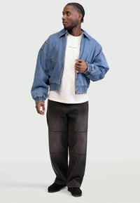 Man wearing a light blue denim jacket over a white t-shirt and black wide-leg pants, looking to his left with one hand adjusting his jacket.