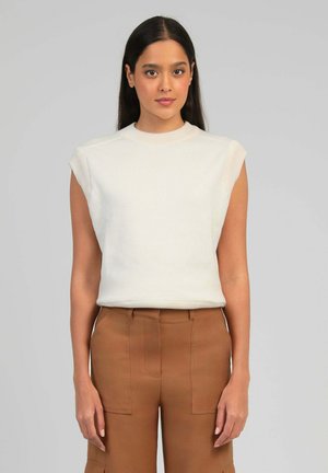 Femme aux cheveux longs et foncés portant un haut blanc sans manches et un pantalon marron taille haute, debout devant un fond gris clair uni.