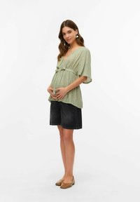 Zelena cvetlična bluzica z V-izrezom in nabrano blago na rokavih, povezana s temno denimu kratkimi hlačami in svetlobrumimi balerinkami. Gladko blago, sproščen kroj.